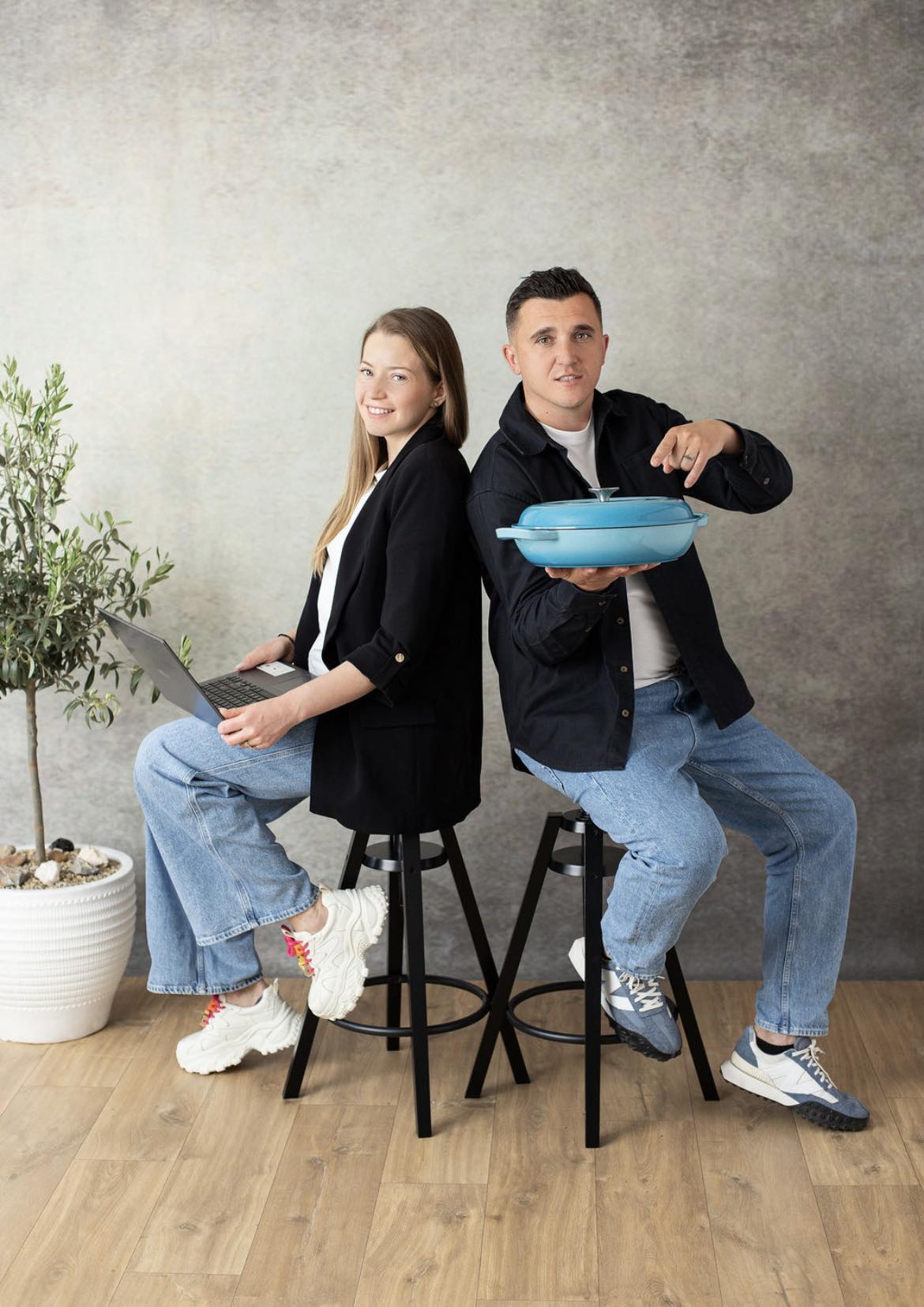 Two people sitting on stools holding a blue pan and a laptop, with a plant in the background.